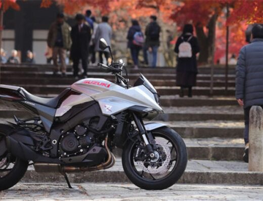 Una moto Suzuki Katana grigia parcheggiata su una strada acciottolata con persone e alberi sullo sfondo in autunno.