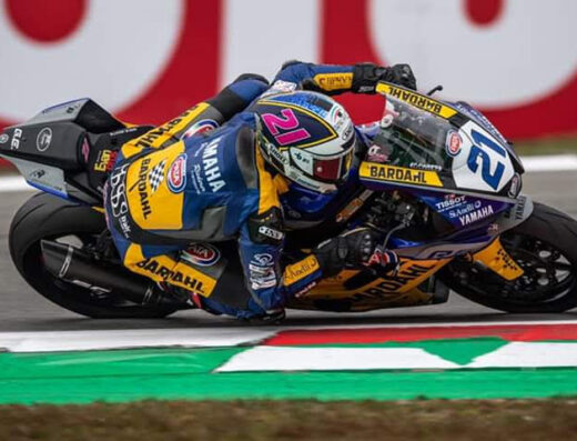 Pilota di moto Yamaha in curva durante una gara, con tuta e casco personalizzati. Sponsor Bardahl ben visibili sulla moto e sull'abbigliamento. Gomma e sgomma in azione.