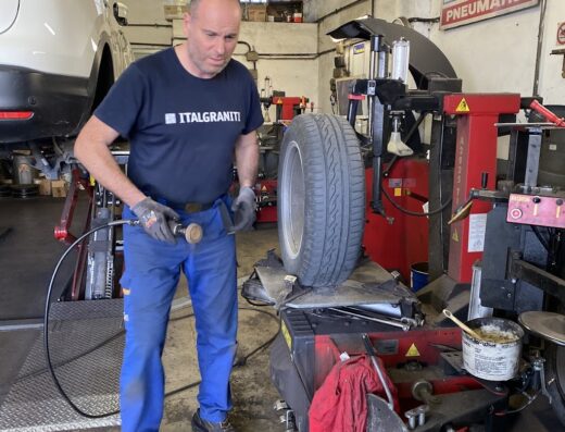 Un gommista al lavoro su un pneumatico Impero in un'officina, con una macchina smontagomme rossa e una gomma pronta per essere riparata o sostituita.