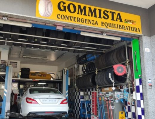 Immagine dell'officina Gommista Roberto V., con una Mercedes bianca sollevata per la convergenza ed equilibratura dei pneumatici. Si vedono anche diversi scaffali di pneumatici.