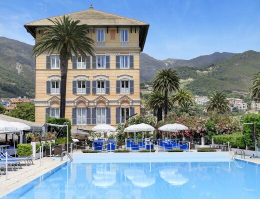 Grand Hotel Arenzano vista piscina con lettini blu e ombrelloni bianchi, palme e sfondo montuoso, in Liguria, Italia