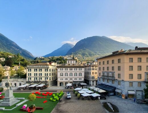 Vista panoramica del Grand Hotel della Posta ad Aosta, con la piazza centrale, i tavolini all'aperto e le montagne sullo sfondo.