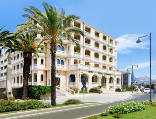 Grand Hotel President Olbia vista esterna con palme, architettura elegante, cielo azzurro e strada antistante. Hotel di lusso a Olbia, Sardegna.