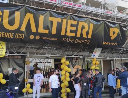 Inaugurazione di Gualtieri Safe Bikers a Rende (CS), negozio di abbigliamento e accessori moto. Persone davanti all'entrata, con banner e palloncini gialli. Scooter e moto parcheggiate. Rivenditore Rev'it.
