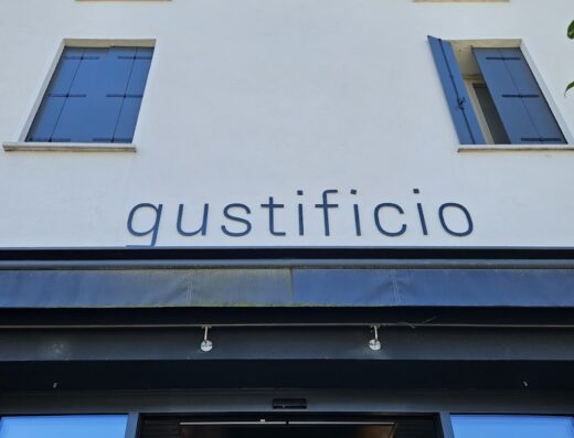 Facciata del Gustificio, negozio con insegna ben visibile e finestre blu. Architettura tipica italiana, design minimale.