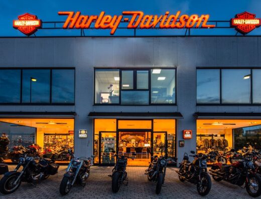 Concessionaria Harley-Davidson illuminata di sera, con motociclette in esposizione, insegna al neon e vetrine che mostrano accessori e abbigliamento moto.