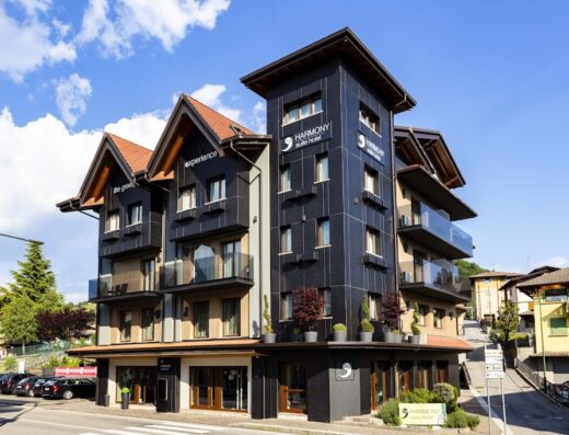 Esterno dell'Harmony Suite Hotel, un edificio moderno con finiture in legno e balconi in vetro, situato in una zona tranquilla con cielo azzurro.