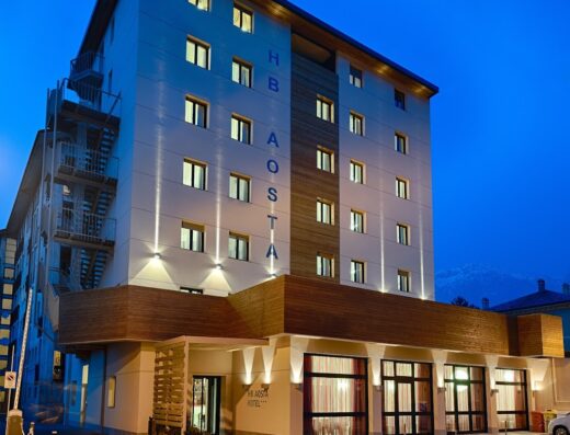 HB Aosta Hotel: Facciata esterna dell'hotel HB Aosta con parcheggio, illuminazione notturna e vista sulle montagne circostanti.