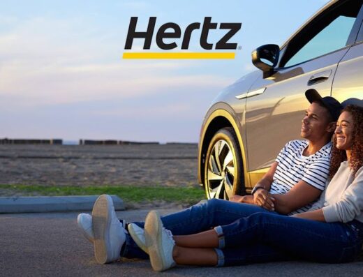 A couple relaxes at sunset near a Hertz rental car, enjoying their scenic road trip.