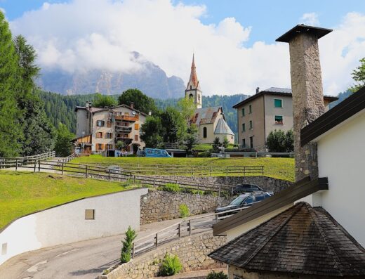 Vista panoramica dell'Hg Hotel La Caminatha a Zoldo Alto, con edifici tradizionali, una chiesa con guglia e le Dolomiti sullo sfondo.