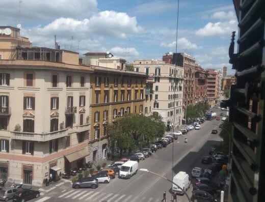 Vista di una strada di Roma dal Home 57 Relais GuestHouse, con auto parcheggiate, persone che attraversano la strada e cielo nuvoloso.