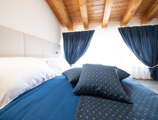 Camera da letto di hotel accogliente con letto matrimoniale, cuscini blu decorativi, soffitto con travi in legno a vista e tende blu che incorniciano la finestra.