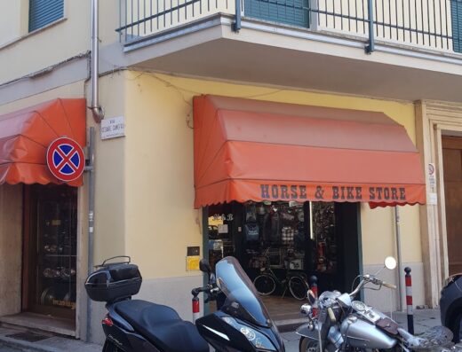 Foto della facciata del negozio Horse and Bike Store di Cristina Bagliani con moto e scooter parcheggiati davanti, negozio di accessori e marketplace per moto e viaggi.