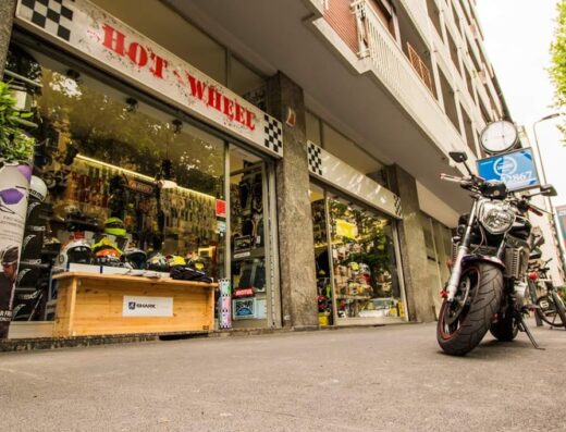 Negozio di accessori per moto "Hot Wheel" con vetrine piene di caschi, protezioni e abbigliamento tecnico. Una moto è parcheggiata di fronte.