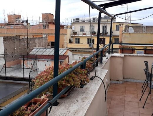 Vista panoramica dalla terrazza di un hotel a Roma, Italia, con tavoli, sedie e vasi di fiori. Edifici storici e antenne sullo sfondo.