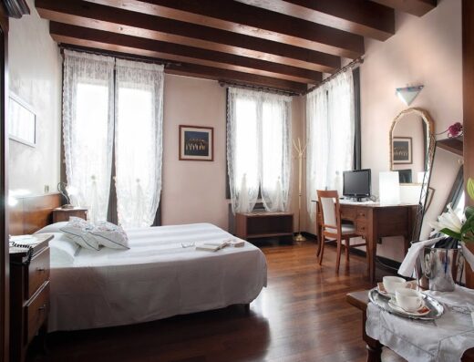 Camera accogliente all'Hotel Al Castello con letto matrimoniale, scrivania in legno, finestre con tende di pizzo e travi a vista, ideale per un soggiorno confortevole.