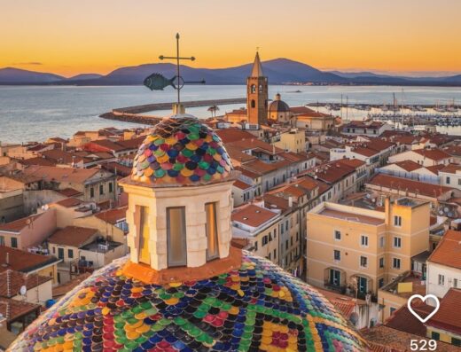 Alghero, Italy: View from Hotel Angedras featuring a colorful tiled dome, cityscape, and the Mediterranean Sea at sunset.