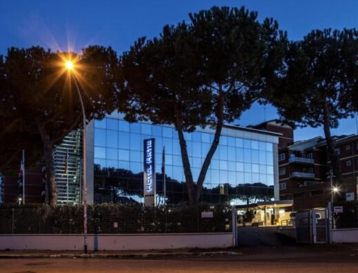 Esterno dell'Hotel Artis al crepuscolo, con facciata a specchio illuminata, insegna luminosa verticale e pini marittimi che incorniciano l'edificio. Atmosfera accogliente e moderna per viaggiatori.