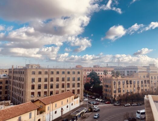 Vista aerea di Roma con cielo azzurro e nuvole bianche, palazzi storici e traffico urbano, Hotel Artromano sullo sfondo.