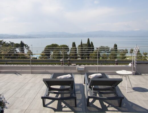 Vista panoramica dal Hotel Berta: due lettini prendisole con asciugamani bianchi su una terrazza con vista sul lago e le montagne, ideale per il relax e il turismo.