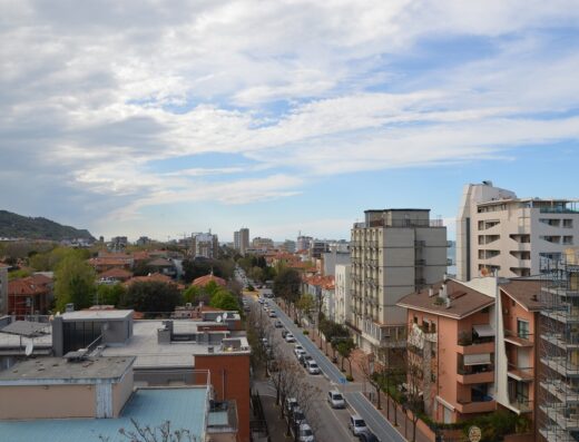 Vista panoramica di Viale Cesare Battisti a Riccione, con edifici residenziali, auto parcheggiate e sfondo collinare sotto un cielo nuvoloso.