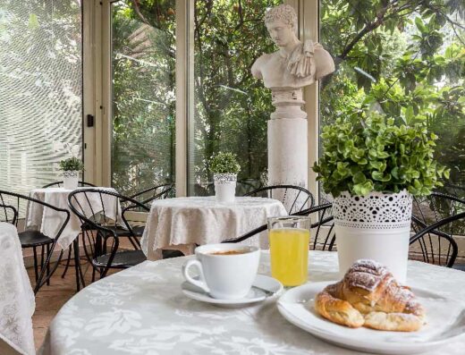 Interno di hotel con tavoli apparecchiati per la colazione, croissant e bevande, statua decorativa sullo sfondo e vista sul giardino