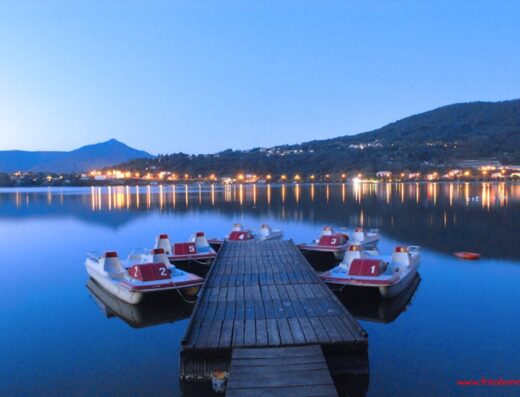 Lago di Avigliana al crepuscolo con pedalò ormeggiati a un pontile di legno. Luci della città riflesse nell'acqua.