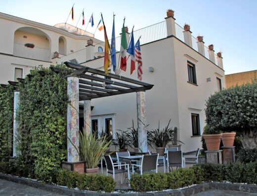 Vista dell'Hotel Costa con bandiere di diverse nazioni, tra cui Italia e Stati Uniti, che sventolano sul tetto. Tavoli e sedie all'aperto invitano al relax.
