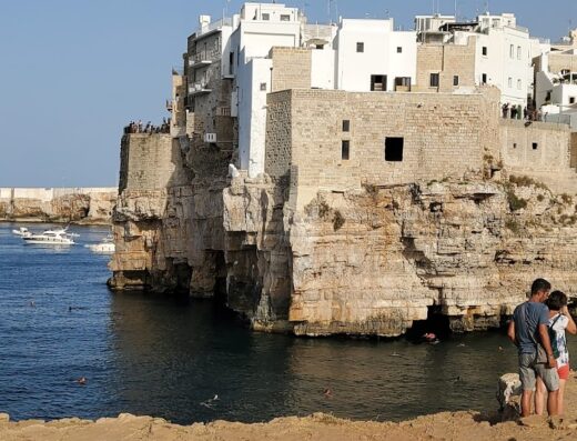 Veduta panoramica dell'Hotel Covo dei Saraceni a Polignano a Mare, con scogliera a picco sul mare, architettura tipica pugliese e turisti che ammirano il paesaggio.