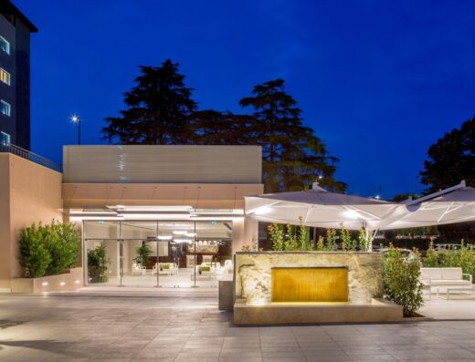 Hotel Cristoforo Colombo entrance at dusk with illuminated fountain, outdoor seating under umbrellas, and glass doors leading inside.