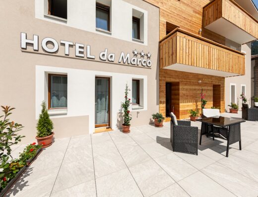 Esterno dell'Hotel Da Marco con tavoli e sedie in vimini sulla terrazza pavimentata, ingresso e balconi in legno chiaro.