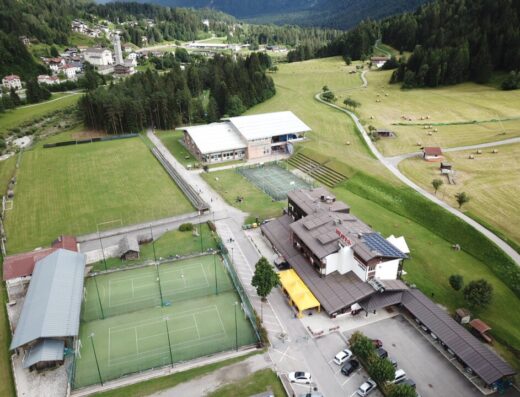 Vista aerea dell'Hotel Davòst con campi da tennis, campi da gioco e parcheggio auto, immerso nel paesaggio montano.