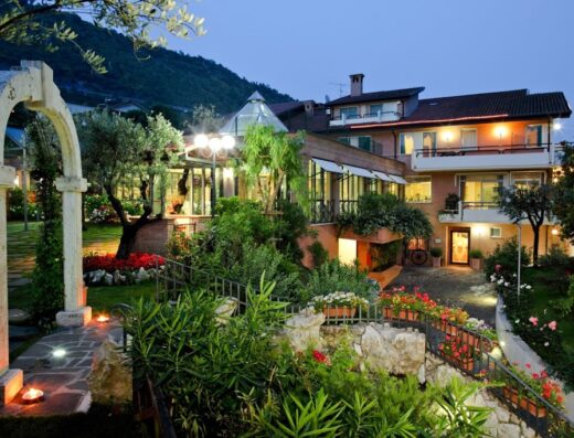 Exterior view of Hotel Des Reves Poggio di Casalucense at dusk, showcasing the lush garden, stone archway, and illuminated building facade.