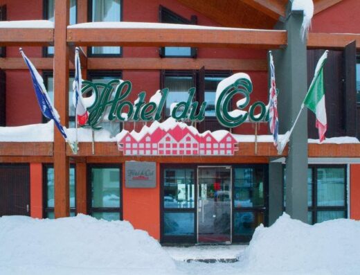 Vista frontale dell'Hôtel Du Col in inverno, con neve, bandiere francese, inglese e italiana e l'insegna dell'hotel