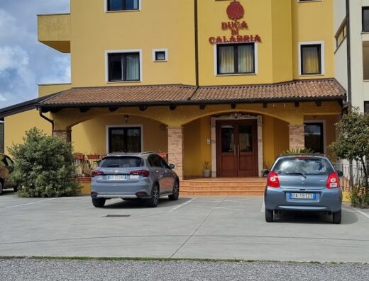 Hotel Duca di Calabria, a yellow building with brown trim, featuring two parked cars in front under a partly cloudy sky.