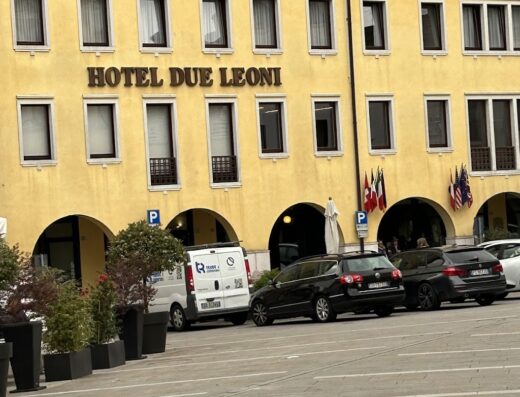 Hotel Due Leoni a Oderzo, Italia, con architettura tradizionale, facciata gialla, finestre simmetriche e auto parcheggiate di fronte. Bandiere nazionali e insegna Hotel Due Leoni visibili.