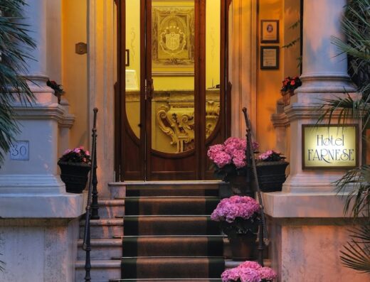 Ingresso principale dell'Hotel Farnese a Roma, con scalinata, tappeto verde con logo, vasi di fiori rosa e insegna luminosa 'Hotel Farnese'.