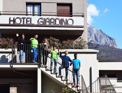 Hotel Giardino Breno: gruppo di persone sorridenti saluta dalle scale esterne dell'hotel con vista sulle montagne circostanti.