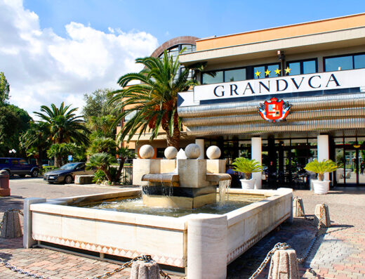 Hotel Granduca Grosseto: vista esterna dell'hotel con fontana decorativa, palme e insegna "GRANDVCA" per un'esperienza di viaggio indimenticabile.