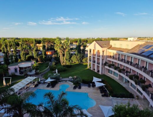 Vista aerea dell'Hotel Isola Sacra con piscina, giardino curato, palme, e edificio rosa con balconi e pannelli solari sul tetto.