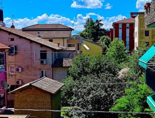 Vista di tetti di case e palazzi in Italia sotto un cielo azzurro con nuvole bianche, vegetazione rigogliosa, atmosfera estiva.