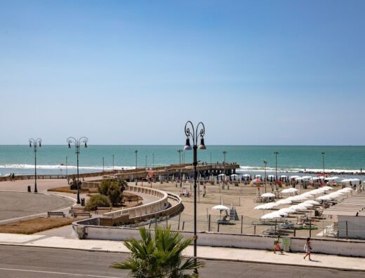 Vista panoramica della spiaggia di Ostia, Roma, con ombrelloni bianchi, stabilimenti balneari, un pontile e il mare azzurro. Hotel La Scaletta Roma