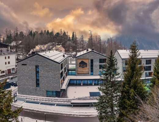 Vista esterna dell'Hotel La Vetta in inverno, con facciata in pietra e neve sui tetti, circondato da alberi e montagne.