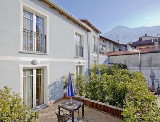 Hotel Lago di Como: Vista esterna dell'hotel con tavolino e ombrellone in terrazza, vegetazione curata e montagne sullo sfondo.