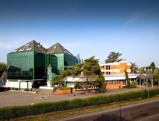 Hotel & Loisirs "Le Ruote": Vista esterna dell'hotel con architettura moderna, parcheggio, aree verdi e segnaletica visibile, ideale per motociclisti e viaggiatori in cerca di comfort e servizi.