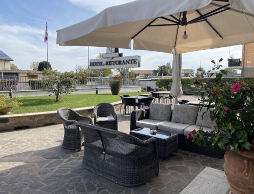 Outdoor patio seating area at Hotel Massimino, featuring wicker furniture, a large umbrella, and a view of the hotel sign and landscaped grounds.