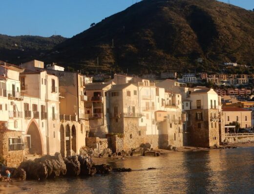 Vista sul lungomare di Cefalù con edifici storici costruiti sulla costa rocciosa, mare calmo e montagna sullo sfondo.