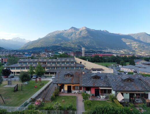 Vista panoramica dell'Hotel Miage con le montagne sullo sfondo, che mostra gli edifici, il parco giochi e la città in lontananza.