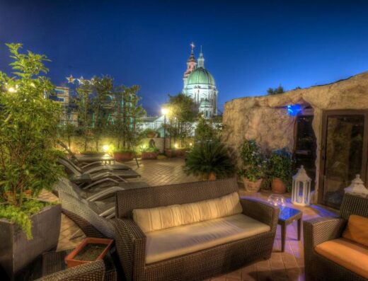 Veduta notturna dell'Hotel Palma: terrazza panoramica con divani in vimini, lettini prendisole, vegetazione e cupola illuminata sullo sfondo.