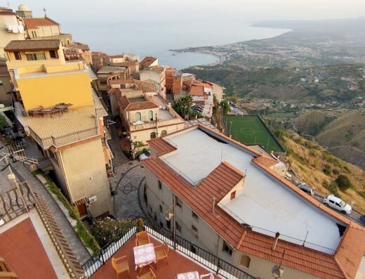 Vista panoramica dell'Hotel Panorama di Sicilia che mostra le stradine acciottolate, gli edifici colorati, il mare in lontananza e un campo da calcio in collina.